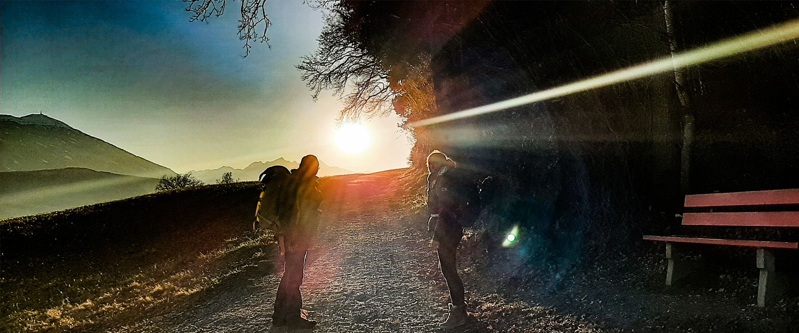 Eine stimmungsvolle Panorama-Aufnahme zweier Wanderer bei Sonnenuntergang. Das warme Licht der tiefstehenden Sonne bricht sich in der Kameraoptik, während im Hintergrund ein markanter Berggipfel und eine rote Ruhebank zu sehen sind.