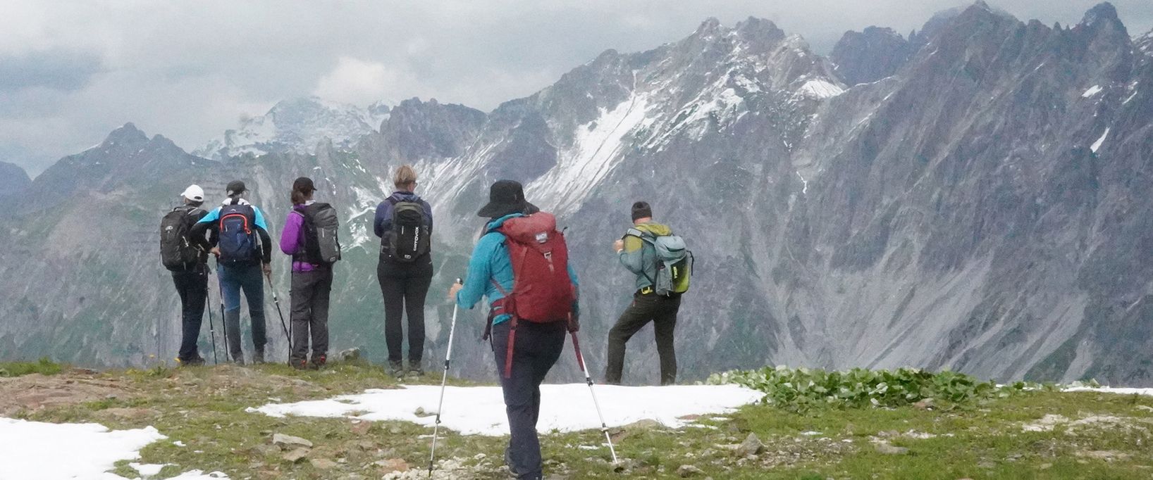 Eine Gruppe von Wanderern mit Rucksäcken und Wanderstöcken steht auf einem Berggipfel und blickt auf ein beeindruckendes Panorama schneebedeckter, schroffer Alpengipfel unter einem bewölkten Himmel.