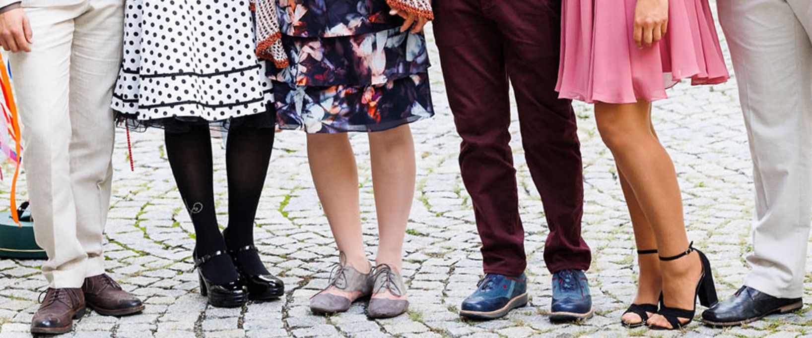 Eine Nahaufnahme der Beine und Schuhe von fünf Personen, die nebeneinander auf Kopfsteinpflaster stehen.