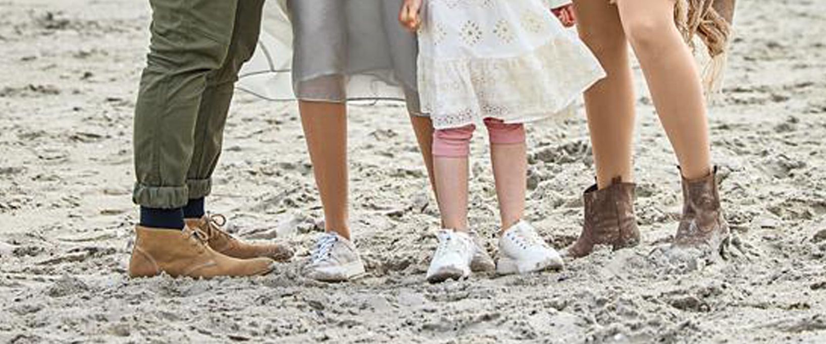 Beine und Füße einer Familie in Freizeitkleidung und verschiedenen Schuhen auf einem sandigen Untergrund.