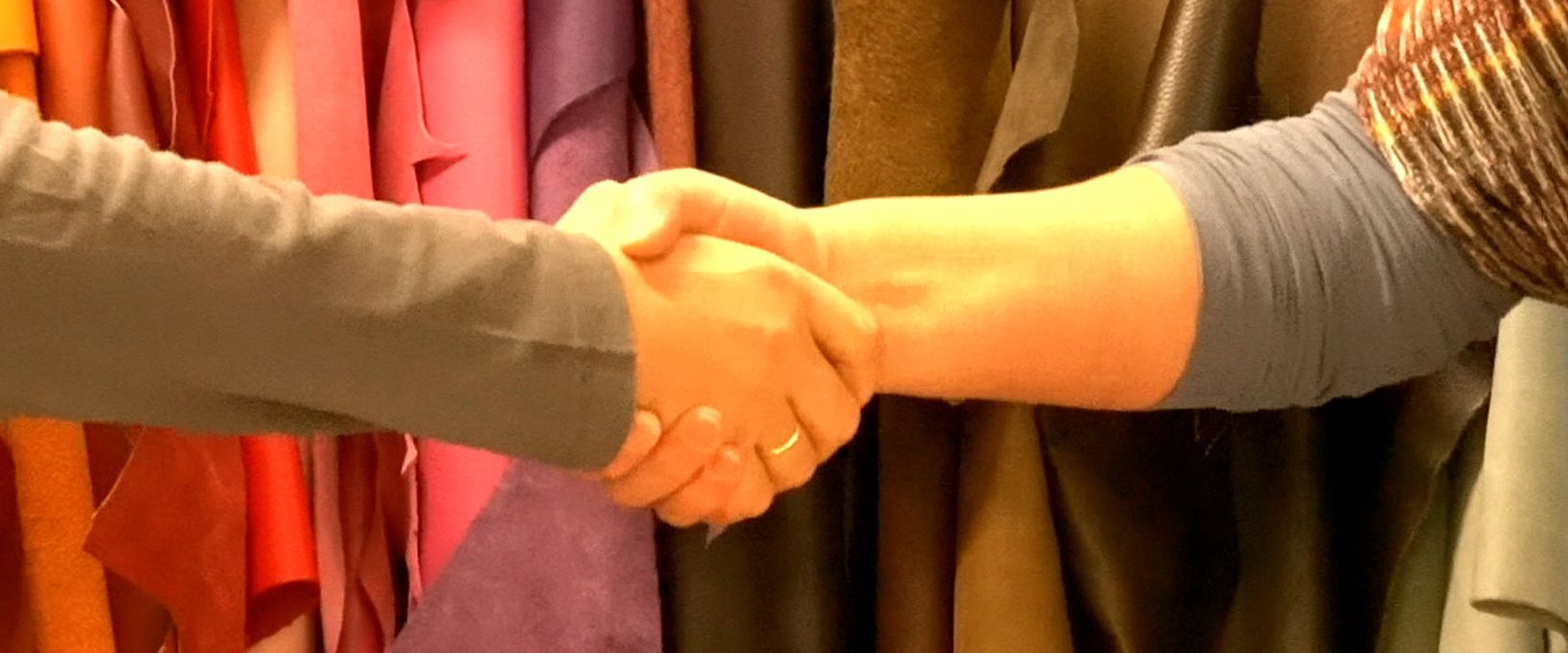 Eine Nahaufnahme eines festen Händedrucks zwischen zwei Personen vor einem Hintergrund aus verschiedenfarbigen Lederrollen, was Vertrauen und partnerschaftliche Zusammenarbeit im Handwerk symbolisiert.